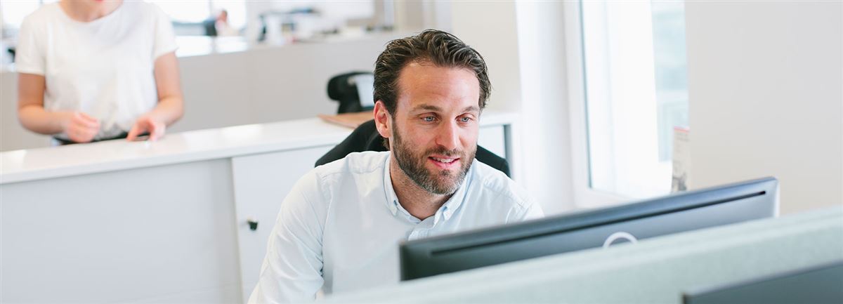 Smiling man in office sitting at a desk behind a computer screen.